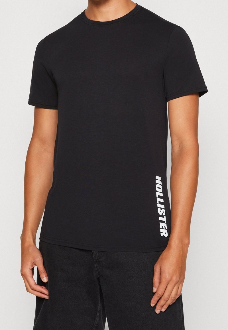 Černé bavlněné tričko s kulatým výstřihem, krátkými rukávy a bílým logem "HOLLISTER" tištěným svisle na levé straně.