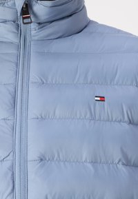 Lys blå pufferjakke med høy krage, vertikal sømdesign og en liten logo-patch med røde, hvite og blå striper.