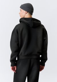 Sweat à capuche noir avec une coupe décontractée, des poignets côtelés et une poche kangourou. Porté avec un pantalon noir assorti et un bonnet en maille gris.