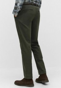 Man som bär olivgröna raka byxor, rutiga skjortan stoppad i byxorna, och bruna mocka anklar stövlar, står med händerna i fickorna.