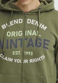 Sudadera con cremallera color verde olivo hecha de tejido suave, con texto en blanco y azul marino: "BLEND DENIM ORIGINAL VINTAGE RECLAMA TUS DERECHOS."