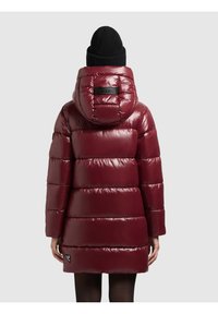 Abrigo acolchado burdeos con capucha, con segmentos acolchados brillantes y un parche con el logo en la parte trasera. Combinado con un gorro negro de punto.