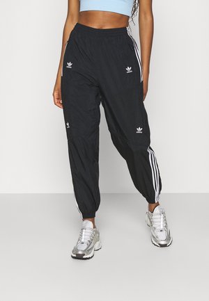 Kobieta ubrana w czarne spodnie dresowe Adidas z białymi paskami i logo, jasnoniebieski top z odsłoniętym brzuchem i srebrne sneakersy, stojąca na białym tle.