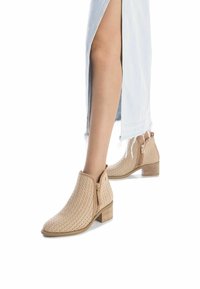 Botas de tobillo beige con una textura tejida, un tacón bajo cuadrado y cremalleras laterales; combinadas con una falda de mezclilla clara con una abertura.