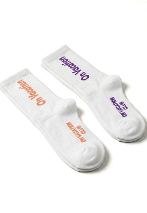 Paire de chaussettes blanches montantes avec texture côtelée, l'une avec un texte orange "On Vacation" et l'autre avec un texte violet "On Vacation Club".