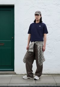 Marinblå t-shirt, rutig skjorta knuten runt midjan, bruna byxor med smala ränder, vita sneakers, grå keps, ståendes mot en grön dörr.