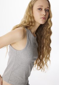 Giovane donna con lunghi capelli biondi ondulati, che indossa un top grigio a coste senza maniche, in posa contro uno sfondo bianco.