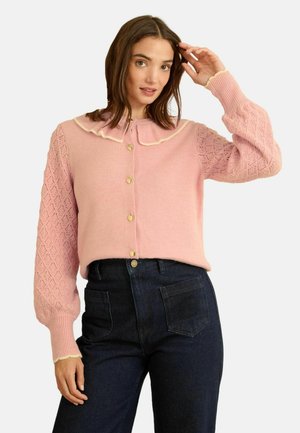 Pink button-up cardigan with a ruffled collar, textured diamond-patterned sleeves, and ribbed cuffs, paired with dark denim trousers.