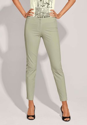 Vrouw draagt lichtgroene slim-fit broek gecombineerd met zwarte open teen hakken, staand met handen in de heup tegen een effen achtergrond.