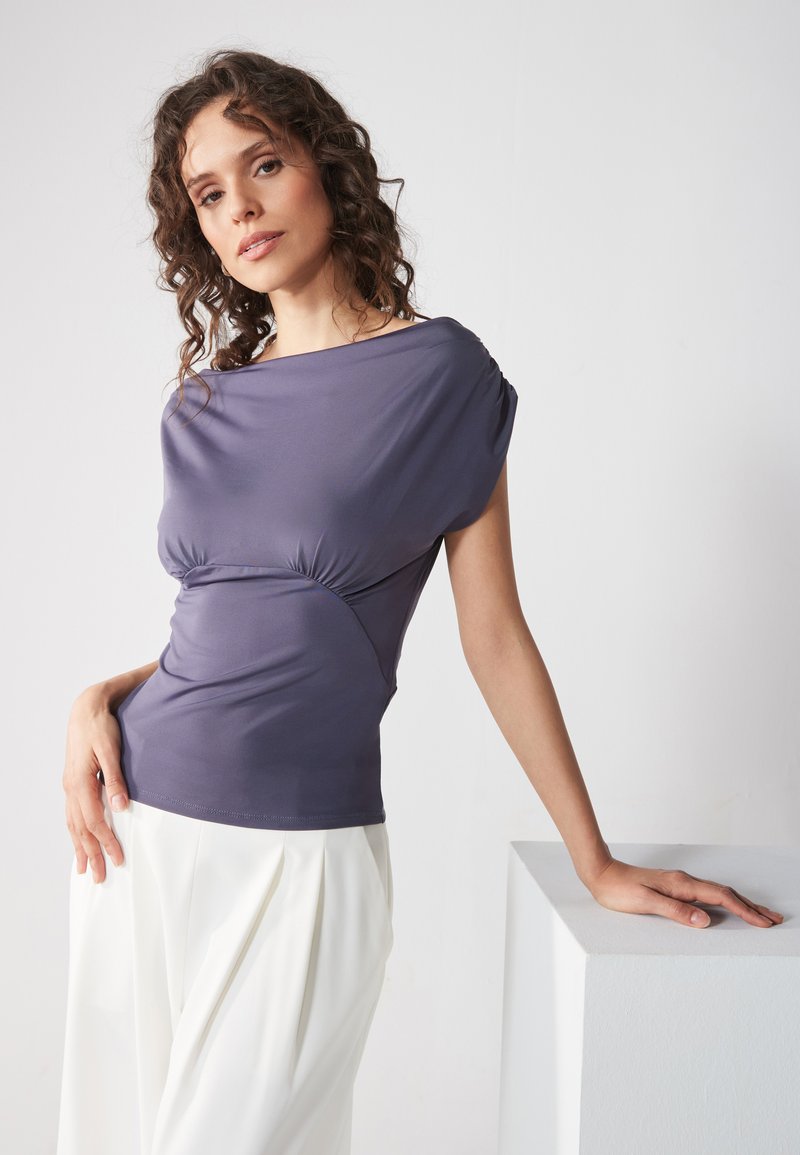Haut ajusté violet avec un décolleté drapé et un détail sur le côté froncé, fabriqué dans un tissu lisse. Associé à un pantalon blanc plissé.