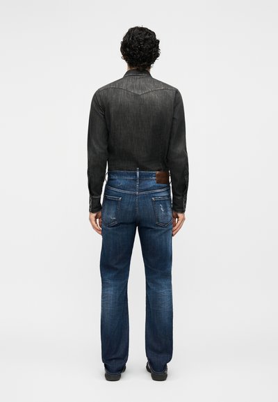 Homme aux cheveux bouclés, vu de dos, portant une chemise en jean noire et un jean bleu avec des poches arrière visibles et un léger effet usé.