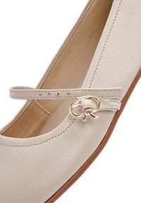 Beige leather Mary Jane flat shoe with a small strap and a gold-tone decorative buckle accent. Smooth texture with rounded toe design.