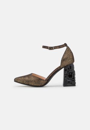 Zapato de punta afilada con parte superior de brillo dorado, correa ajustable en el tobillo y tacón ancho negro texturizado con detalles geométricos.