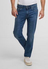 Donkerblauwe denim jeans met een rechte pijp, voorzien van voorzakken, een knoopsluiting en een versleten textuur. Gecombineerd met witte sneakers.