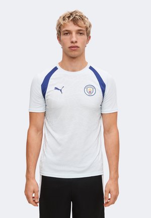 Junger Mann trägt ein weißes Manchester City Sportshirt mit blauen Akzenten und schwarze Hose, steht vor einem einfarbigen Hintergrund.