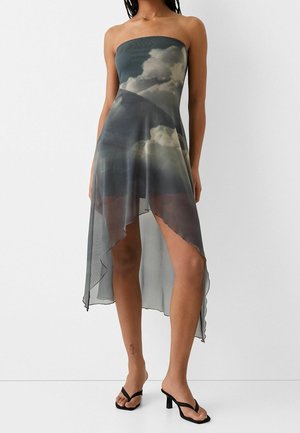 Robe asymétrique bustier avec imprimé nuages, superposition transparente, portée avec des sandales noires à talons hauts, modèle debout sur fond uni.