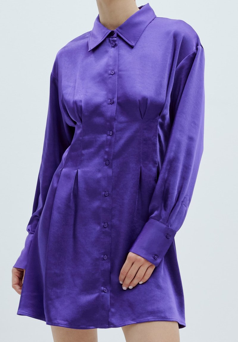 Lila Satinhemdkleid mit langen Ärmeln, knopflosem Vorderteil und geschwungendem Saum. Verfügt über einen klassischen Kragen und Faltendetails an den Seiten.