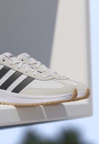 Paar weiße und beige Adidas-Sneaker mit schwarzen Streifen und Gummisohlen, die auf einer weißen Kante vor einem blau-grauen Hintergrund liegen.