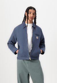 Jachetă din canvas albastru cu guler, închidere cu fermoar și un buzunar pe piept. Asortată cu pantaloni verzi deschis. Textură netedă, design casual.