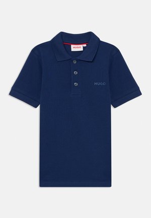 Jūras zila polo krekls, kas izgatavots no gludas kokvilnas. Tam ir klasiskā apkakle, trīs pogu aizdare un smalka logotipa detaļa audumā.