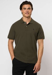 Muž s krátkými tmavými vlasy, oblečený v olivově zeleném polokošili s krátkým rukávem a černých kalhotech, stojí s jednou rukou v kapse na bílém pozadí.