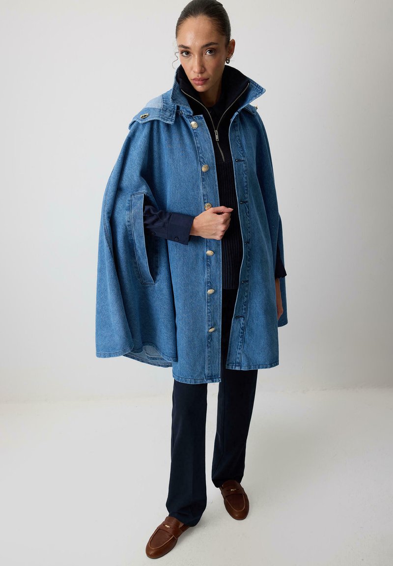 Denim capejakke har en høj krave, store knapper og sidelommer. Stoffarven er lyseblå med en glat tekstur.