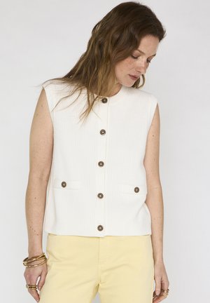 Donna con capelli castani ondulati indossa un gilet bianco senza maniche abbottonato e pantaloni giallo pallido, guarda verso il basso con le braccia rilassate lungo i fianchi.