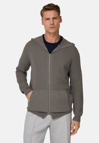 Grauer Reißverschluss-Hoodie mit Kapuze, gerippten Bündchen, Fronttaschen und glatter Textur. Wird über einem marineblauen Hemd getragen, kombiniert mit hellen Hosen.