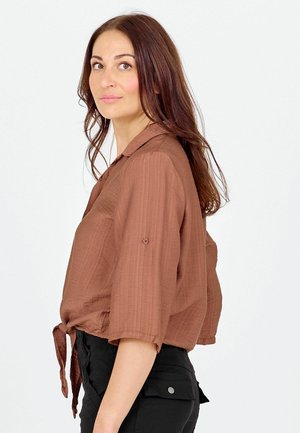 Sieviete ar gariem brūniem matiem, valkājot vaļēju brūnu kreklu ar pogām un sasietu pie jostas, kā arī melnas bikses, stāvot sāniski pret baltu fonu.