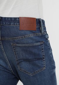 Närbild av en bakficka på blå denimjeans med en brun läderlabel från Superdry på midjan och en vit inskjuten skjorta.