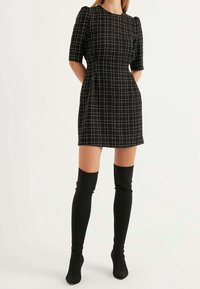 Schwarzes karierte Kleid mit kurzen Puffärmeln, strukturiertem Stoff und figurbetonter Silhouette, kombiniert mit kniehohen schwarzen Stiefeln.