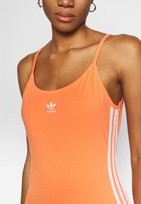 Orange tanktop i bomull, designad med tunna axelband, en rundad halsringning och med vita Adidas-ränder på sidorna.