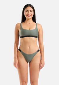 Conjunto de bikini verde oliva con escote cuadrado y tirantes gruesos. La parte superior tiene una banda negra con la marca; la parte inferior ofrece una cobertura mínima.