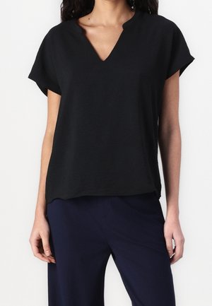 Femme portant un chemisier noir à manches courtes avec un col en V en encoche et un pantalon bleu marine foncé, debout devant un fond clair uni.