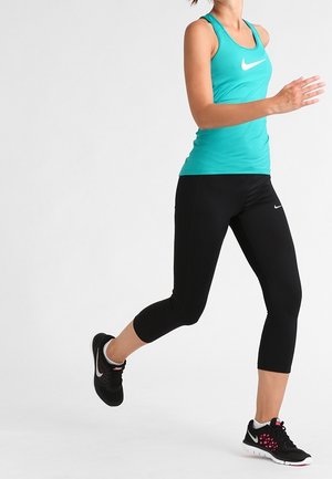 Frau trägt türkisfarbenes Tanktop und schwarze Leggings, läuft im mittleren Schritt vor einem einfarbigen weißen Hintergrund.