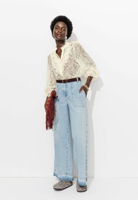 Blouse en dentelle avec des détails froncés, jean large taille haute bleu clair, ceinture marron, sac à frange rouge et chaussures à enfiler imprimé léopard.