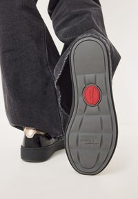 Fekete lakkbőr cipő kerek orral, strukturált szürke gumitalppal, piros kényelmi logóval és kontrasztos fémsarkakkal.