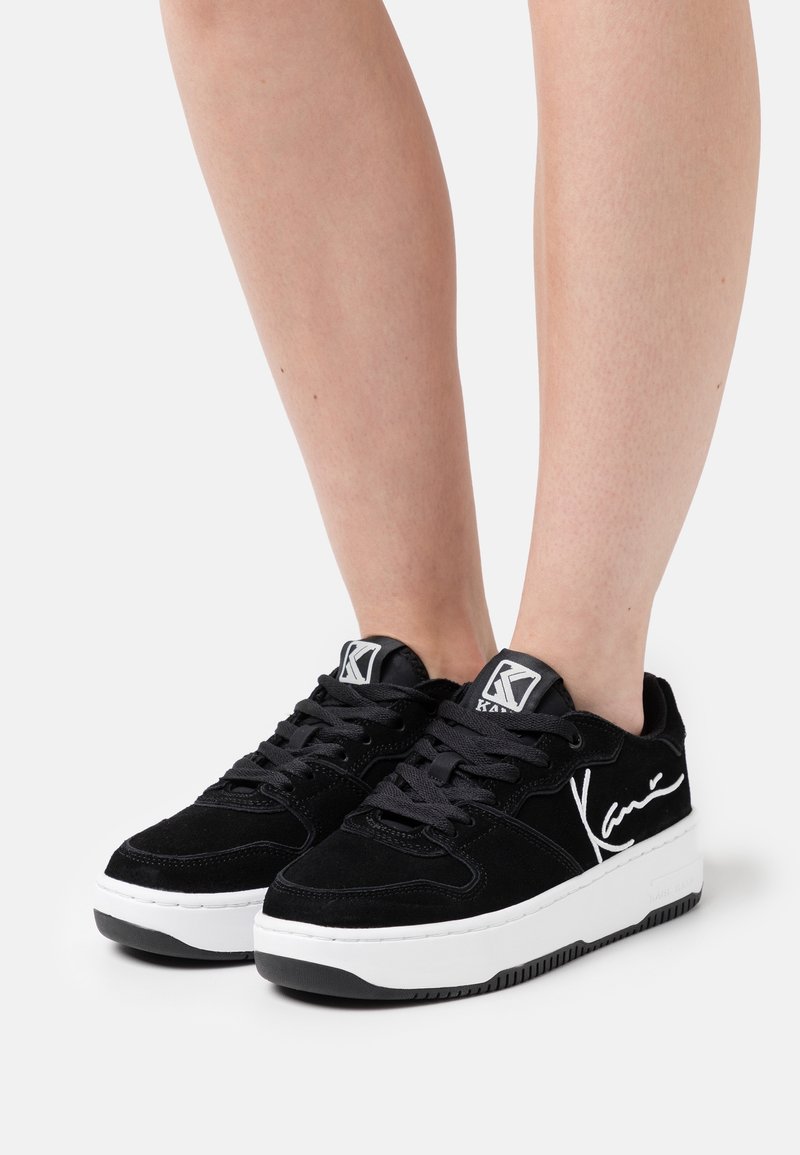 Baskets basses noires avec semelles blanches épaisses, lacets noirs et logo en écriture blanche sur le côté, portées sur jambes nues sur un fond uni.