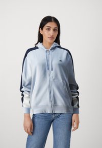 Svetlomodrá mikina na zips s kapucňou na šnúrku, námornícke rukávy, zelené logo na prsiach a predné vrecká; vyrobená z mäkkej látky s hladkou textúrou.