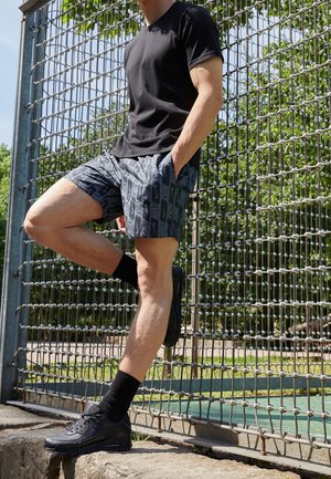 Homme en chemise noire, short à motifs, chaussettes et chaussures noires, appuyé contre une clôture métallique à l'extérieur, une jambe levée et les mains dans les poches.