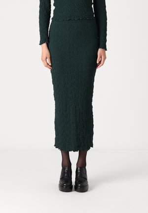 Vrouw draagt een donkergroene, getextureerde lange rok met bijpassende top, zwarte transparante panty's en zwarte platformschoenen, staand op een witte achtergrond.