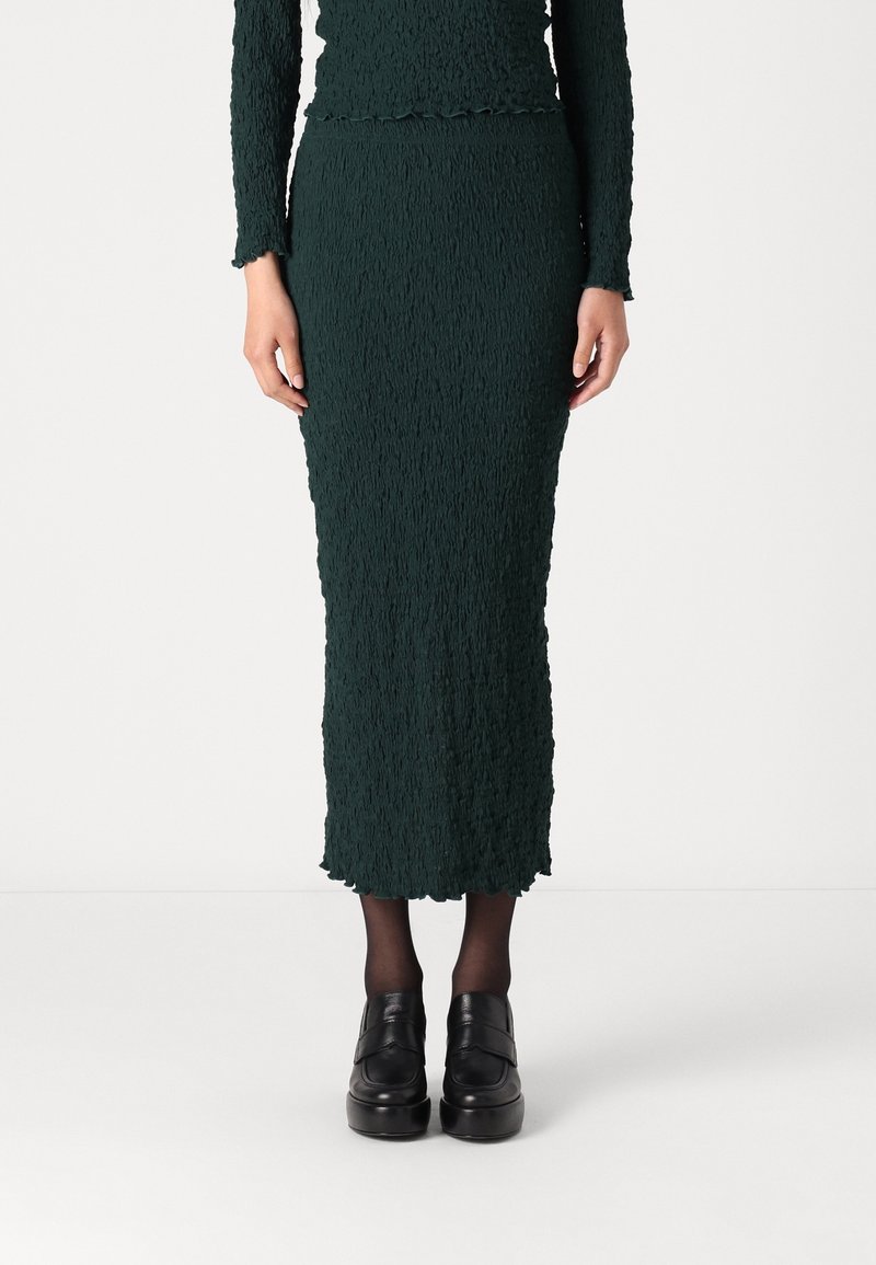 Femme portant une longue jupe texturée vert foncé avec un haut assorti, des collants noirs transparents et des chaussures à plateforme noires, debout sur un fond blanc.