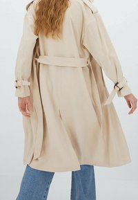 Trench-coat beige avec une taille cintrée, des manches larges et des accents à boucles aux poignets. Texture lisse et design fluide.