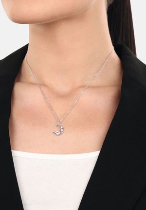 Silberne Halskette mit einem Halbmond-Anhänger, die zwei Verzierungen aufweist, an einer filigranen Kette, die auf einem nackten Hals gegen ein schwarzes Blazer getragen wird.