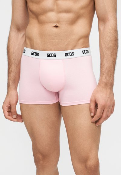 Άνδρας που φορά ανοιχτό ροζ μπόξερ με λευκή ελαστική μέση που φέρει επαναλαμβανόμενο μαύρο λογότυπο GCDS, στέκεται μπροστά σε απλό φόντο.