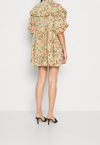 Frau in einem kurzen, lockeren, blumigen Kleid mit Rüschensleeves und schwarzen High-Heels, stehend vor einem schlichten weißen Hintergrund.