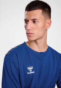 Camicia blu a maniche lunghe realizzata in materiale morbido, con scollatura a V, logo Hummel bianco e dettagli neri testurizzati sulle spalle.