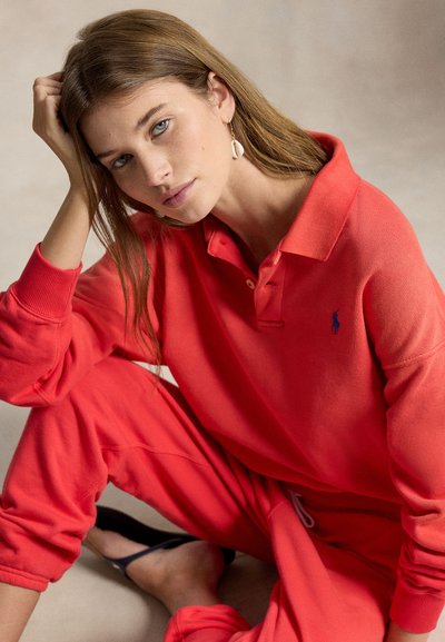 Mujer joven con ojos azules y cabello largo castaño claro, vestida con polo rojo y pantalones a juego, sentada con una mano en el cabello y pendientes de concha visibles.