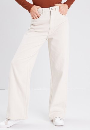 Jeans a gamba larga di colore crema in tessuto denim, con tasche frontali, vita alta e cuciture a contrasto. Abbinati a sneakers bianche.