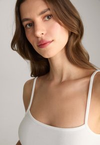 Witte top met dunne bandjes, gladde textuur, ronde halslijn en een aansluitend ontwerp, tegen een neutrale achtergrond.